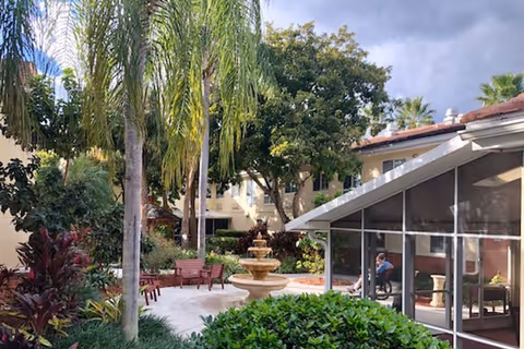 Outdoor courtyard area at Barrington Terrace featuring a multi-tiered stone fountain surrounded by palm trees, bushes, and benches. A screened-in patio area is visible on the right with a person sitting inside. The background shows a multi-story building with windows and a partly cloudy sky.