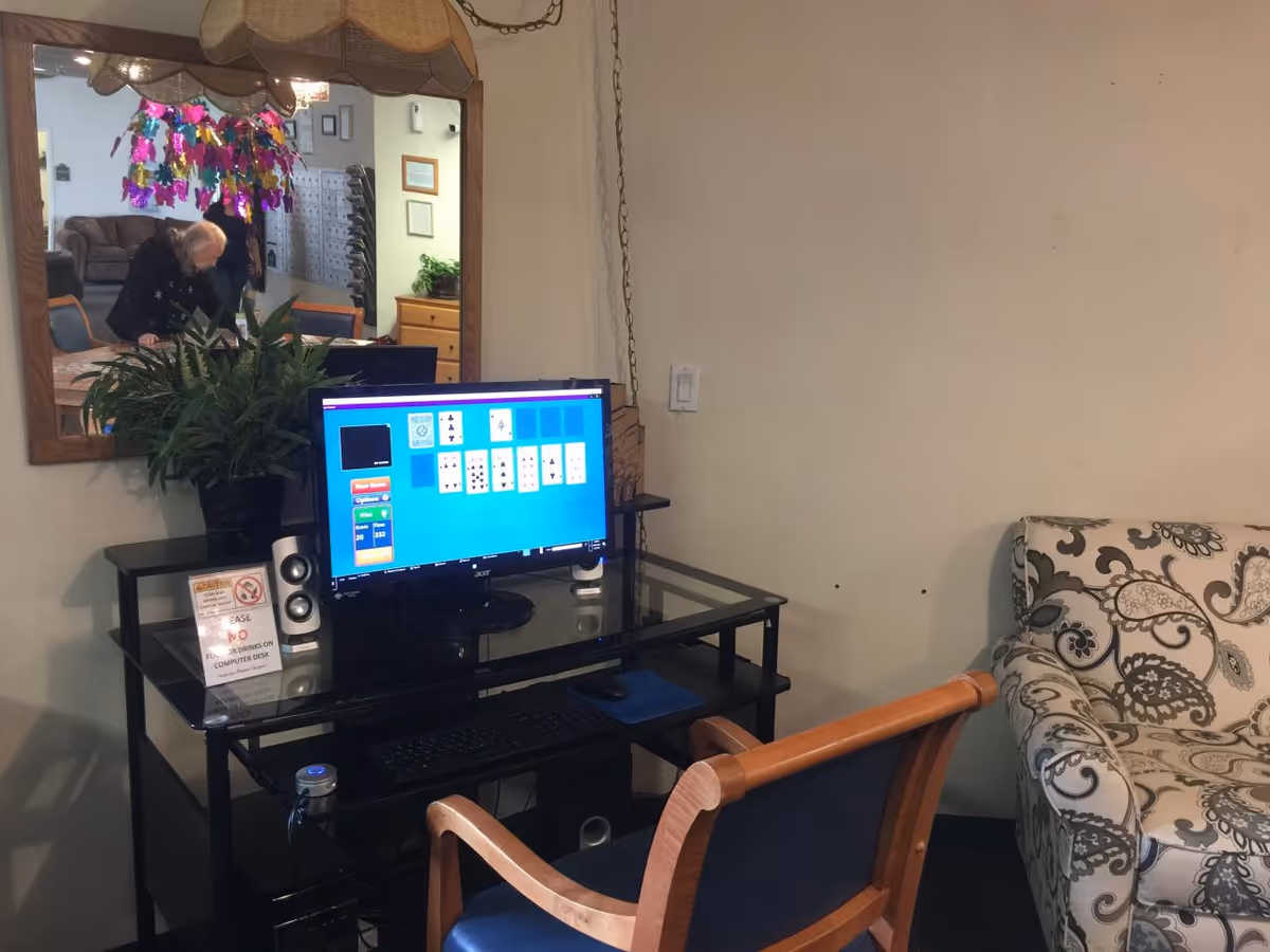 A small computer desk with a monitor displaying a card game, a keyboard, speakers, and a potted plant. A wooden chair with blue upholstery is in front of the desk. To the right is a patterned armchair. A large mirror on the wall reflects a person and part of a room with chairs and a couch.