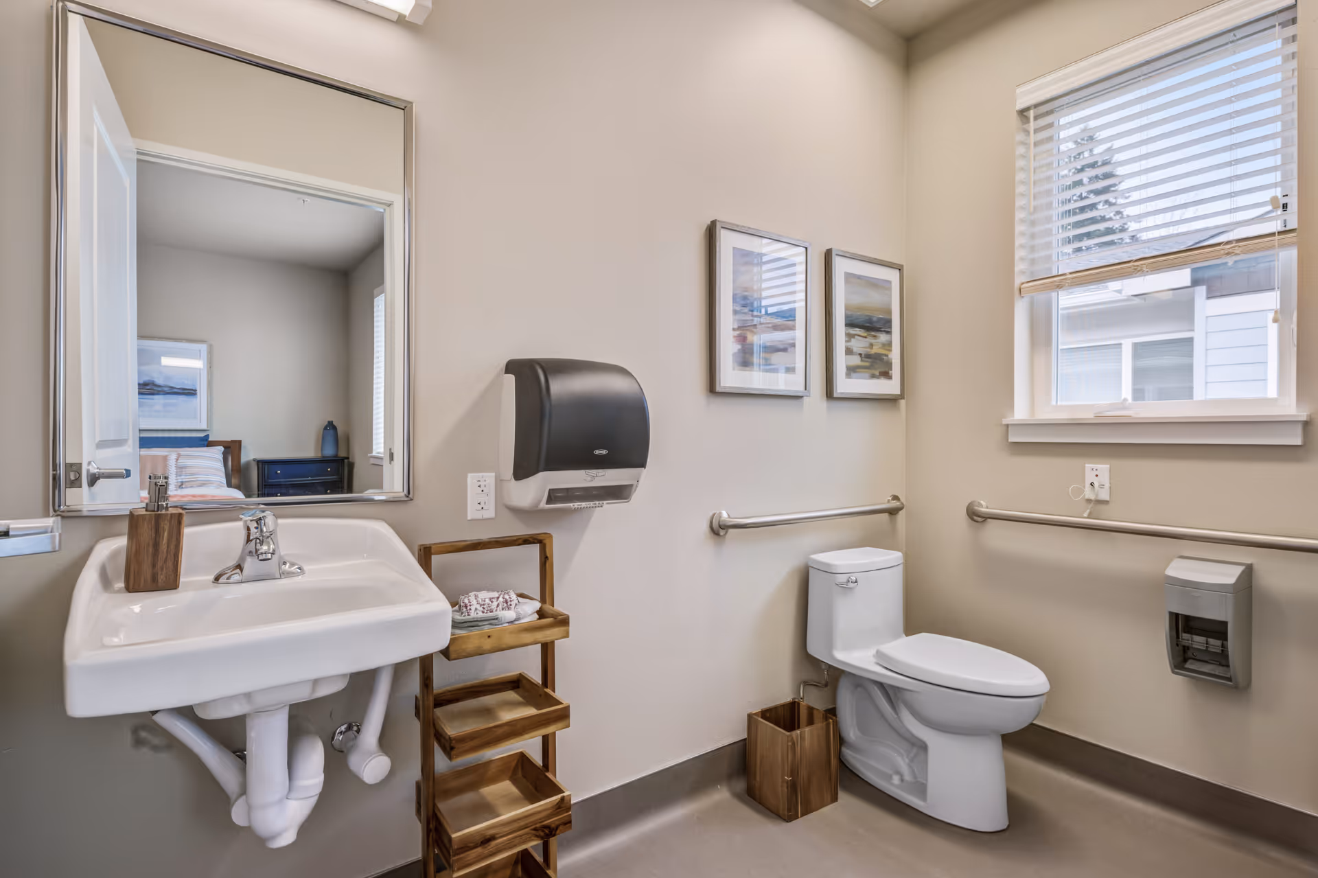 A clean and accessible bathroom with a white sink, a large mirror, a toilet with grab bars on both sides, a wooden storage rack with towels, two framed pictures on the wall, a window with blinds, and a paper towel dispenser mounted on the wall.