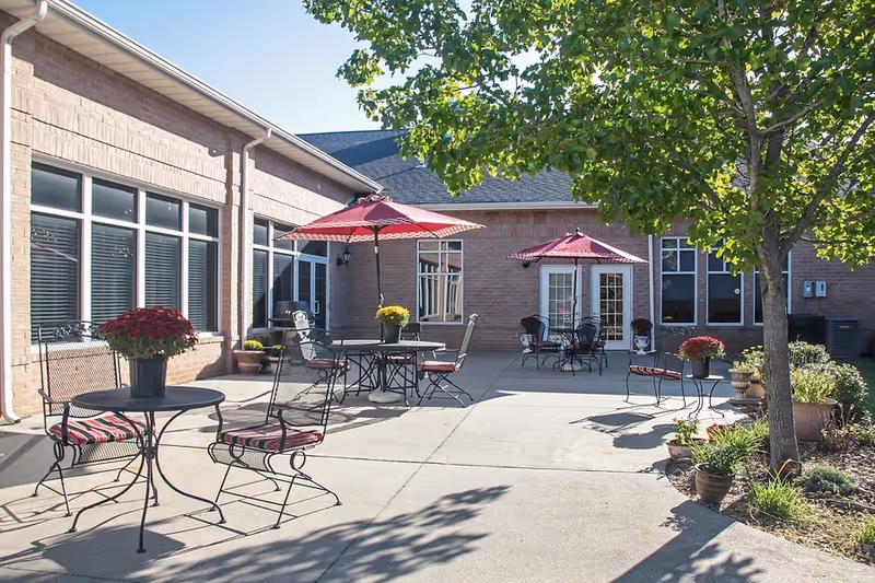 Sunlit outdoor courtyard with metal tables and chairs, red umbrellas, potted plants, and a brick building facade.