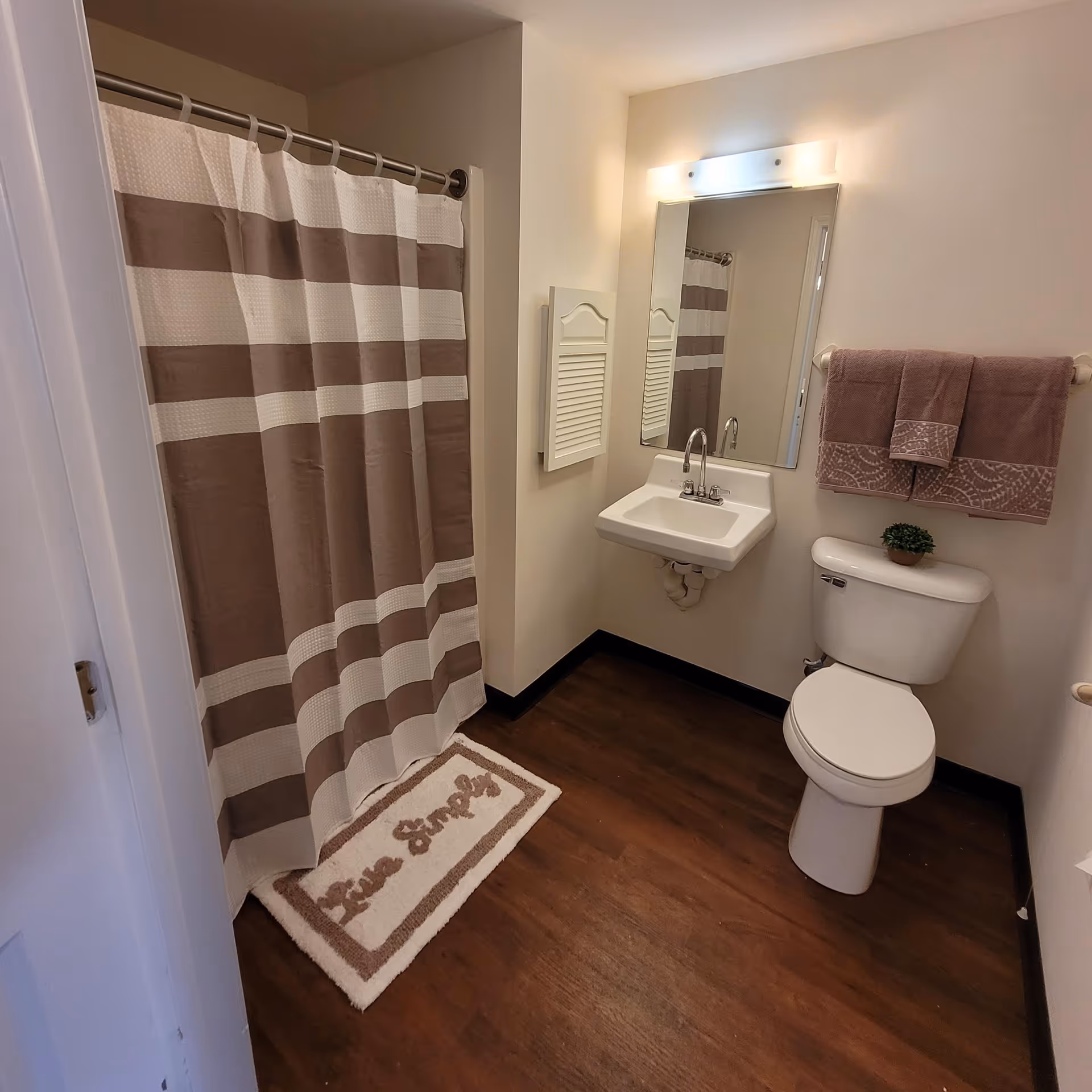 A clean bathroom with a white toilet, a small white sink with a mirror above it, a towel rack holding two brown towels, a shower with a brown and white striped curtain, and a floor mat that says 'Live Simply'. The floor is wooden and the walls are light-colored.