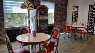 Interior view of a retro-style dining area with round tables and red chairs. There is a vintage jukebox against a brick wall between two windows with blinds. The floor is wooden, and there are decorative wine racks on the walls.