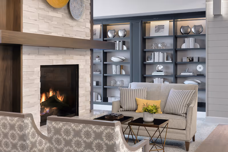 Cozy living room area with a lit fireplace surrounded by light-colored stone. Two patterned armchairs face a beige loveseat with striped and yellow pillows. A small table with a plant and books sits in front of the loveseat. Behind the seating area is a built-in dark gray bookshelf filled with decorative items and books.
