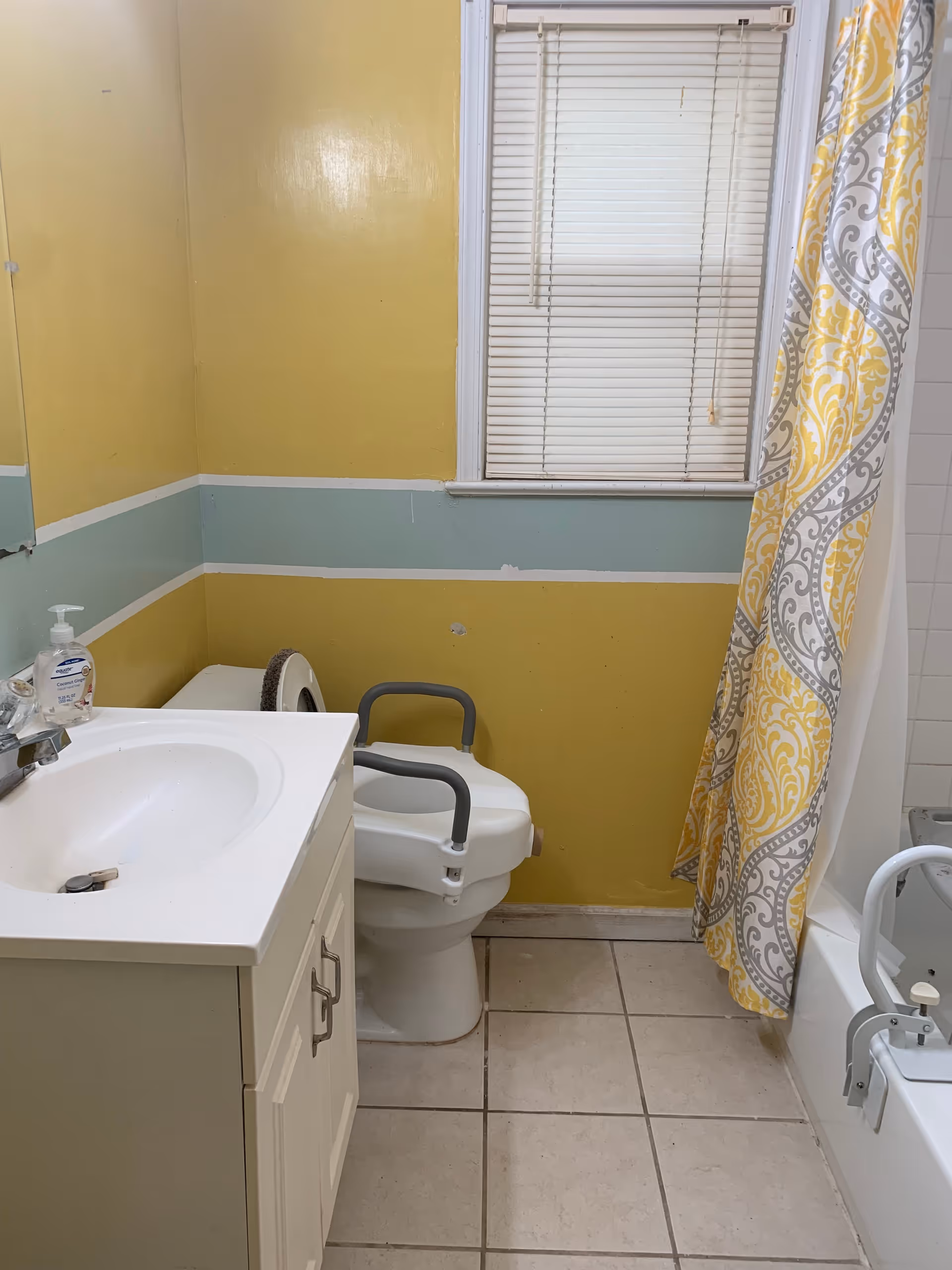 A small bathroom with yellow and light blue painted walls, a white sink with a cabinet, a toilet with safety rails, a window with closed blinds, and a bathtub with a yellow and gray patterned shower curtain and a safety rail attached to the tub.
