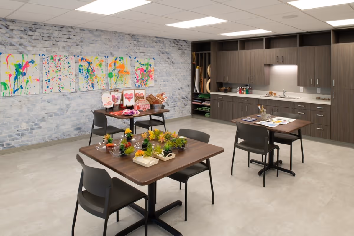 A bright, spacious activity room with light gray brick walls and a light gray floor. The room features three wooden tables with black chairs. One table has small potted plants arranged on it, another has art supplies and coloring pages, and the third has Valentine's Day themed decorations. The back wall has a large built-in cabinet with a countertop, sink, and storage. Colorful abstract paintings are displayed on the wall above the tables.
