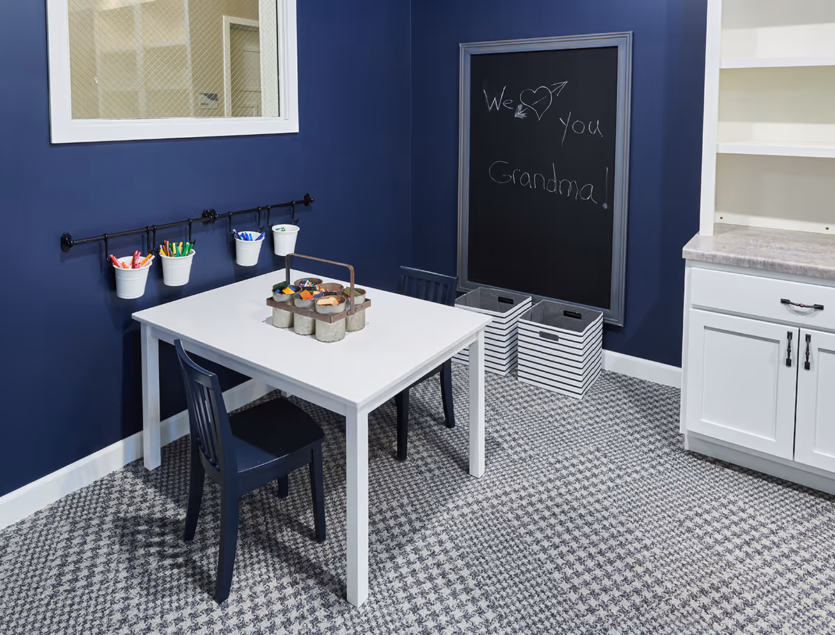 Small craft room with a white table and navy chairs, art supplies hanging on the wall and a chalkboard reading "We ♥ you Grandma!"