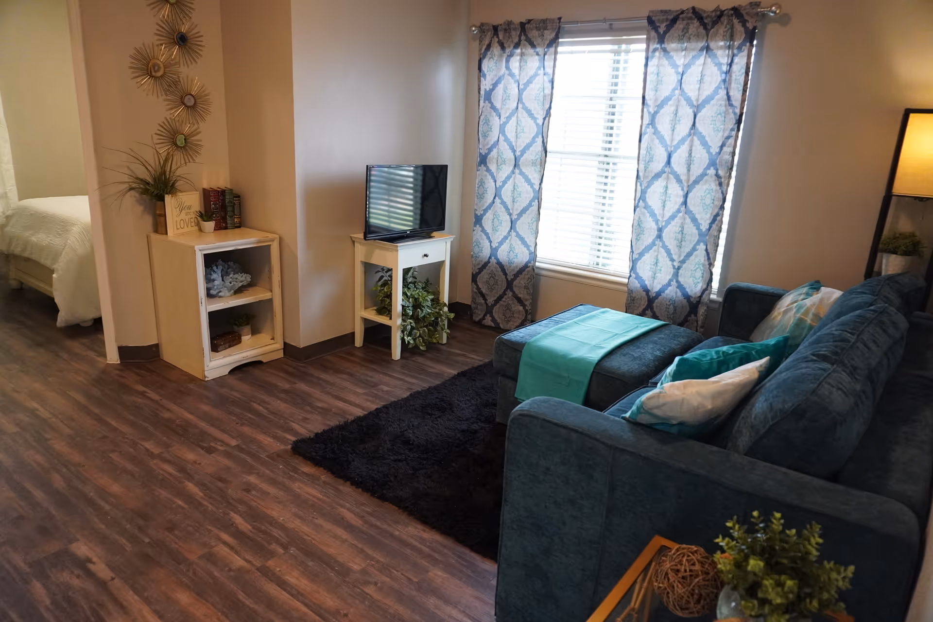 A cozy living room area with a teal sofa adorned with pillows and a matching ottoman with a teal throw. A black rug lies on the wooden floor. A small white table holds a flat-screen TV and a green plant underneath. Blue and white patterned curtains cover a window letting in natural light. To the left, a small white cabinet with decorative items and a sign that reads 'You are Loved' is visible, with a glimpse of a bedroom in the background.