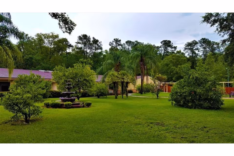 A lush green garden area with various trees and shrubs, including palm trees, surrounding a multi-tiered water fountain. In the background, there is a single-story building with a purple roof partially visible, and a cloudy sky overhead.