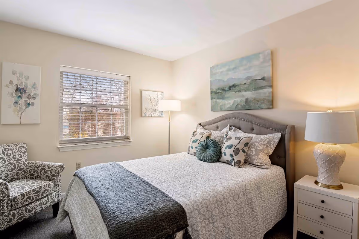 A cozy bedroom with a large bed featuring multiple decorative pillows and a gray upholstered headboard. There is a patterned armchair to the left, a window with blinds, two framed artworks on the walls, and two lamps providing warm lighting, one on a nightstand and one floor lamp in the corner.