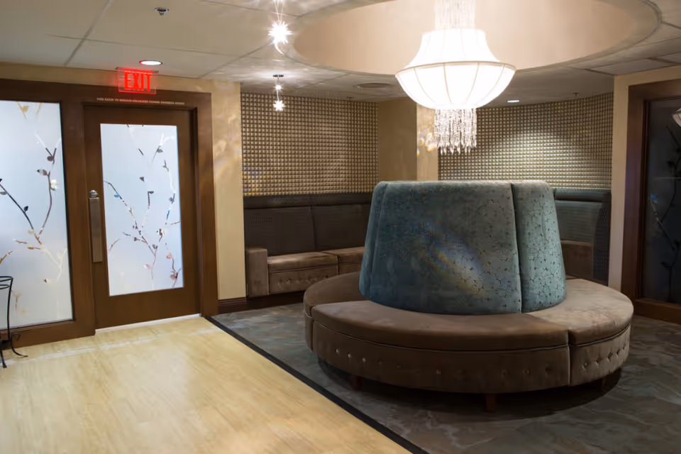 An interior lounge area with a circular cushioned bench in the center, featuring a blue upholstered backrest. The walls have a textured, patterned design, and there is a large chandelier hanging from the ceiling. To the left, there are frosted glass doors with floral designs and an illuminated red exit sign above them.