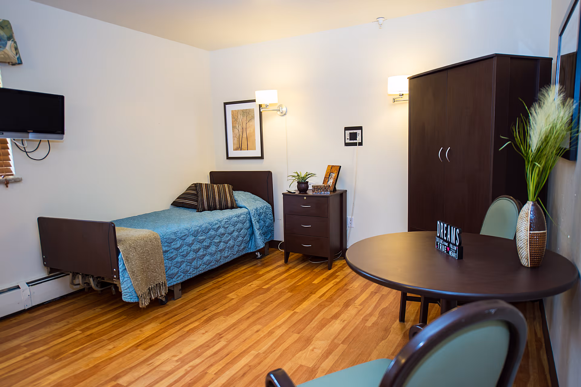 A cozy bedroom in a senior living facility featuring a single bed with a blue quilt and striped pillows, a small dark wooden nightstand with a plant and picture frames, a dark wooden wardrobe, a round table with two chairs, a decorative vase with tall grass, and a wall-mounted TV. The room has wooden flooring and soft lighting from wall sconces.