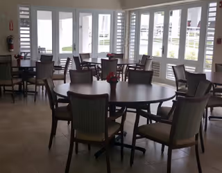 Sunlit dining area with round and rectangular tables and chairs arranged near large windows and glass doors.