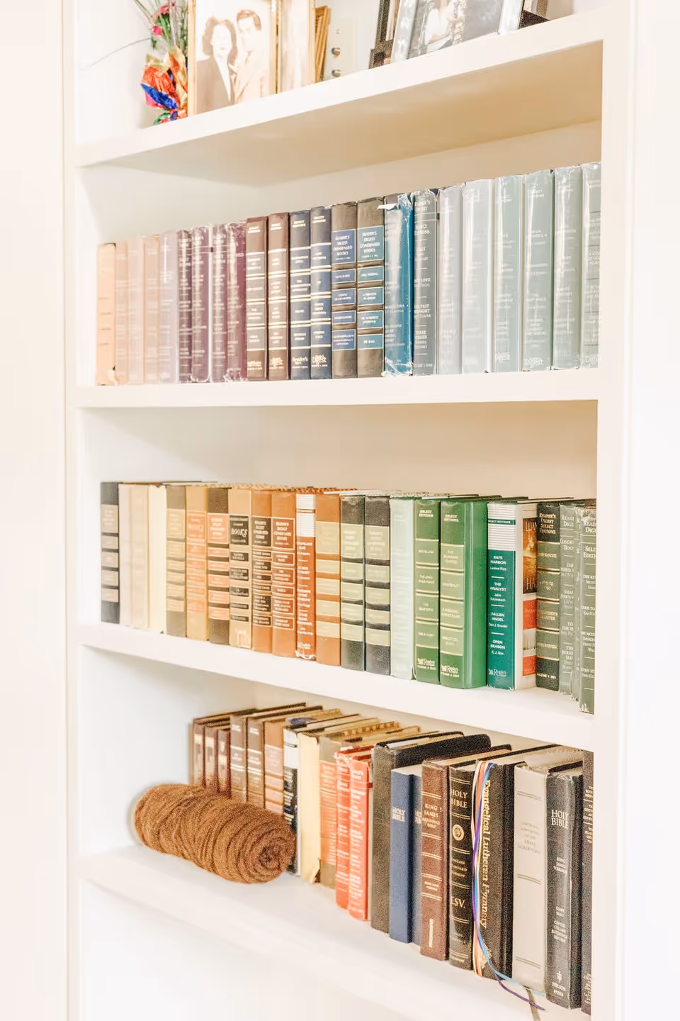 A white bookshelf filled with multiple rows of books, including several volumes of Reader's Digest Condensed Books and various editions of the Holy Bible. On the top shelf, there are framed photographs and a decorative item with colorful ribbons.