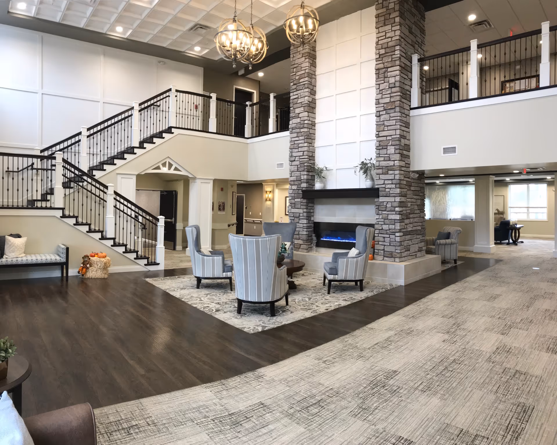 Spacious senior living facility common area with a two-story ceiling, stone fireplace, and modern chandeliers. The room features a seating area with four striped armchairs around a small round table on a patterned rug. There is a staircase with black railings leading to an upper level balcony. The flooring is a combination of dark wood and light carpet, and the walls are painted in neutral tones.