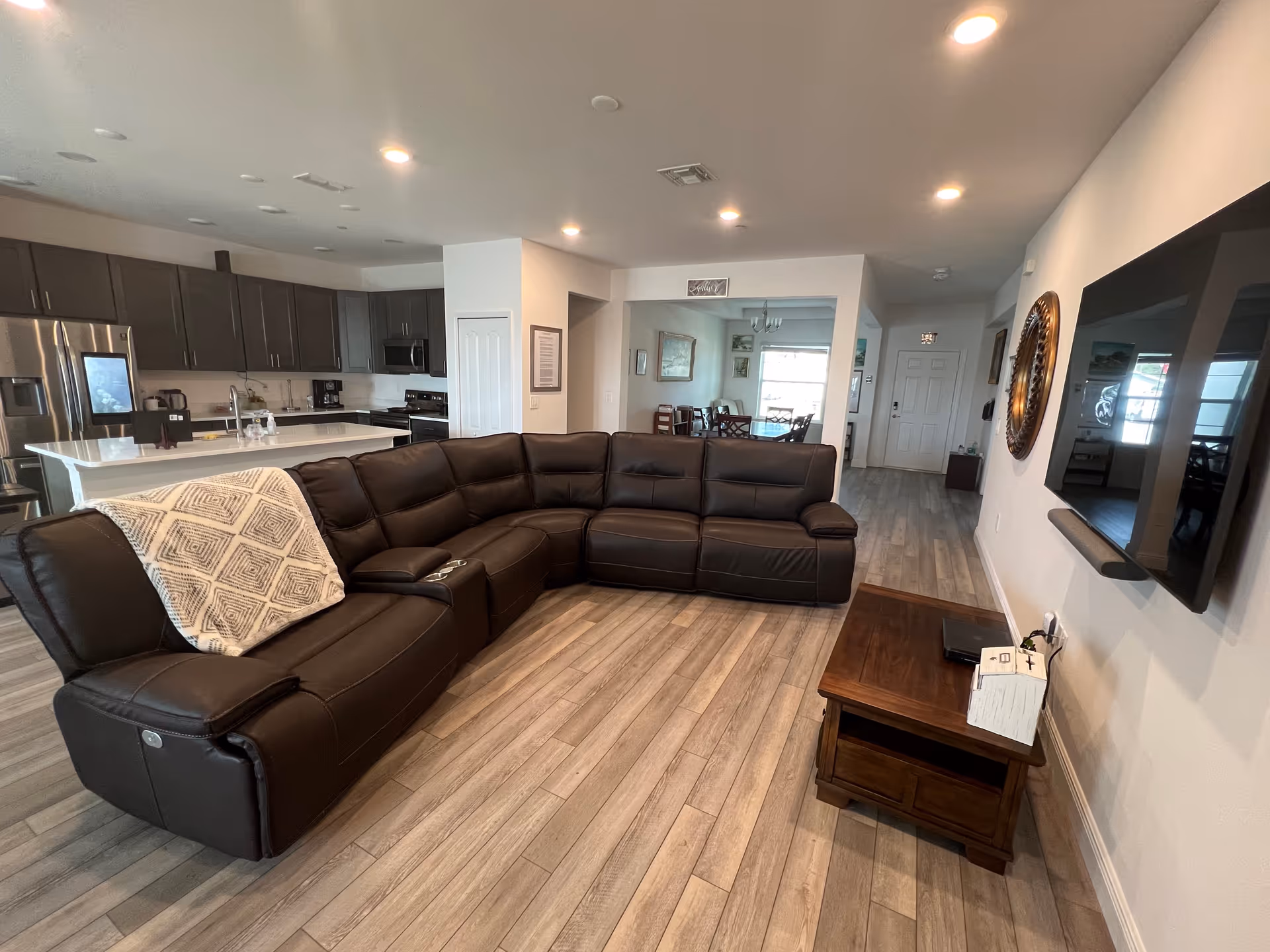 A modern open-concept living area featuring a large dark brown leather sectional sofa with a patterned throw blanket on one side. The room has light wood flooring and recessed ceiling lights. In the background, there is a kitchen with dark cabinetry, stainless steel appliances, and a white countertop island. A wooden side table with a few items on it is placed next to the sofa, and a large flat-screen TV is mounted on the wall opposite the sofa. Further back, a dining area with a table and chairs is visible near a window.