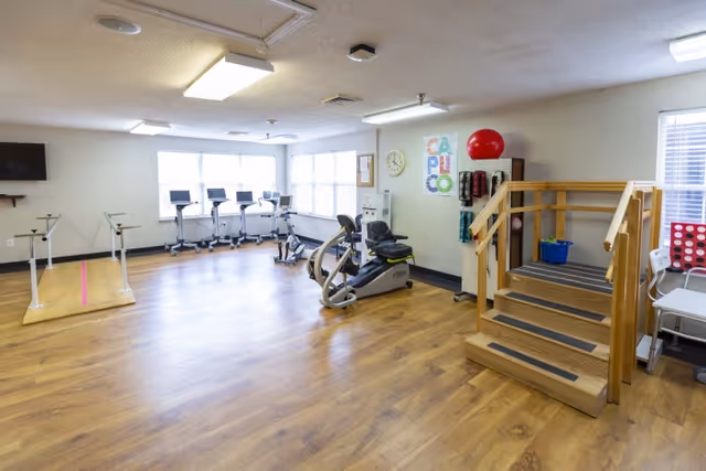 A spacious rehabilitation room with wooden flooring featuring exercise equipment including stationary bikes, parallel bars for walking practice, and a wooden staircase with handrails for physical therapy. The room is well-lit with large windows and has a wall clock and colorful wall art.