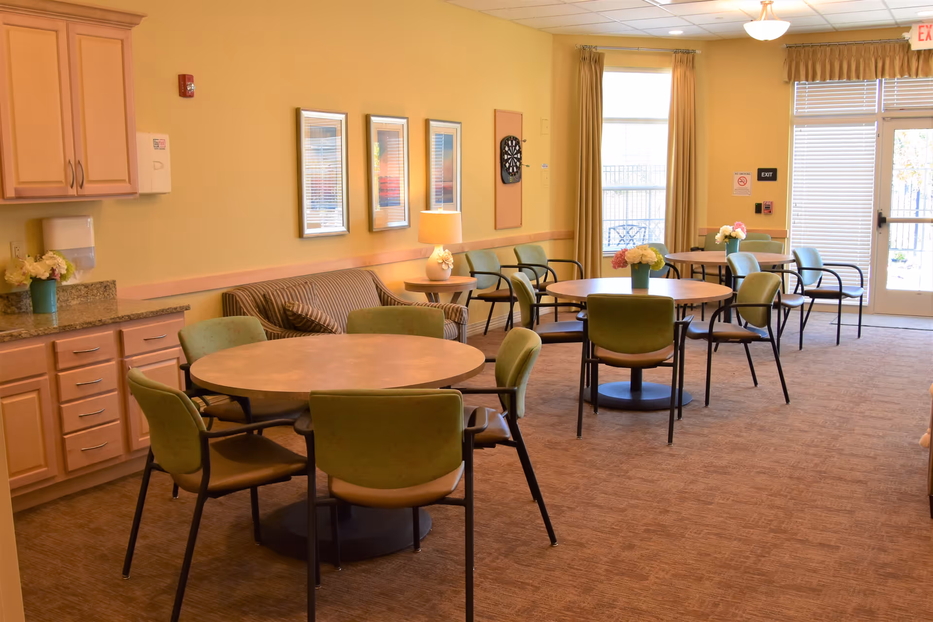 A well-lit common area in an assisted living facility featuring round tables with green chairs arranged around them. There are flower vases on the tables, a couch against the wall, framed pictures, a dartboard, and a lamp on a side table. The room has beige walls, carpeted floor, and large windows with curtains allowing natural light to enter.
