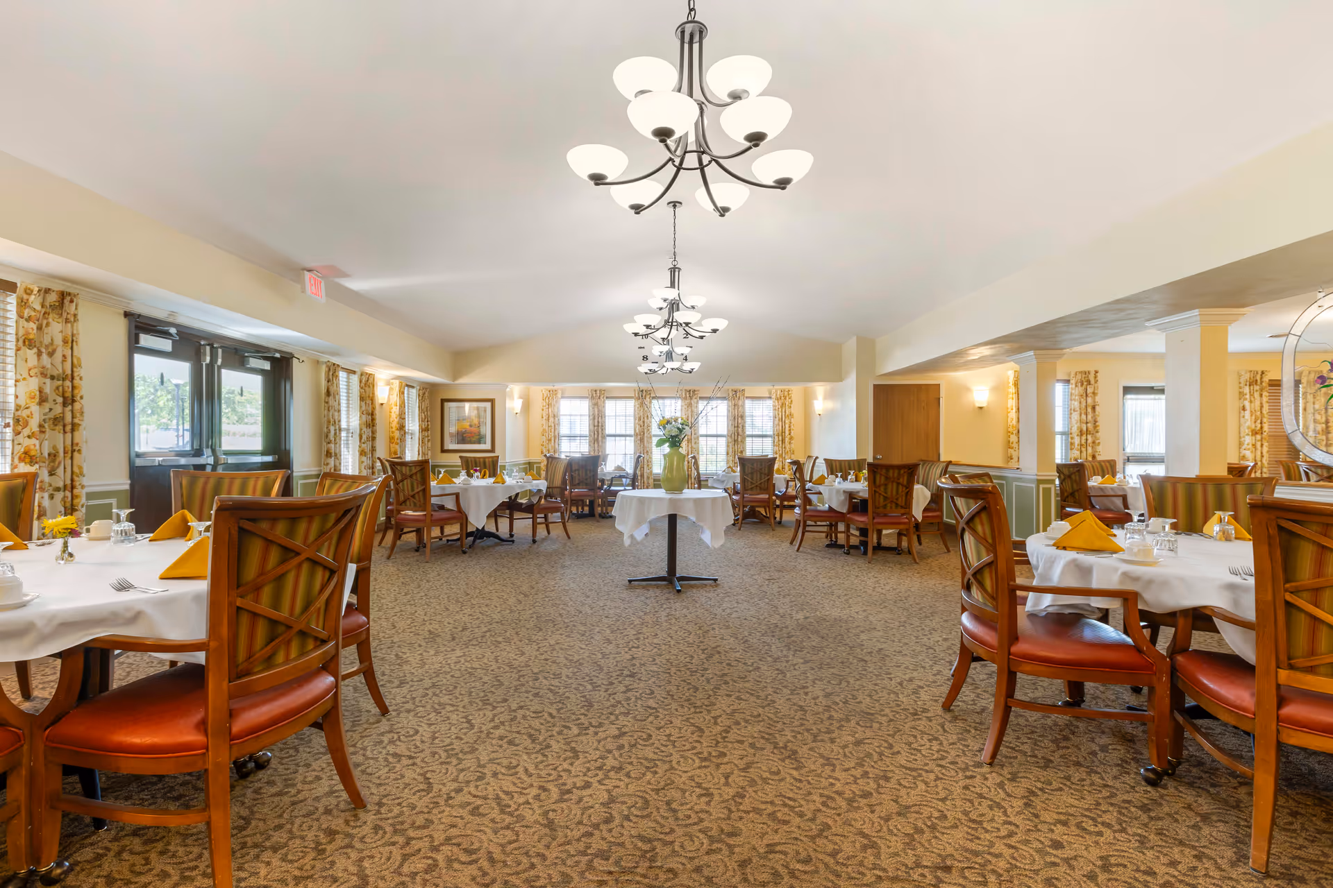 A spacious dining room with multiple round tables covered with white tablecloths and set with yellow napkins, cups, and silverware. The room features patterned carpet, wooden chairs with cushioned seats, large windows with floral curtains, and elegant chandeliers hanging from the ceiling. A vase with flowers is placed on a small round table in the center of the room.