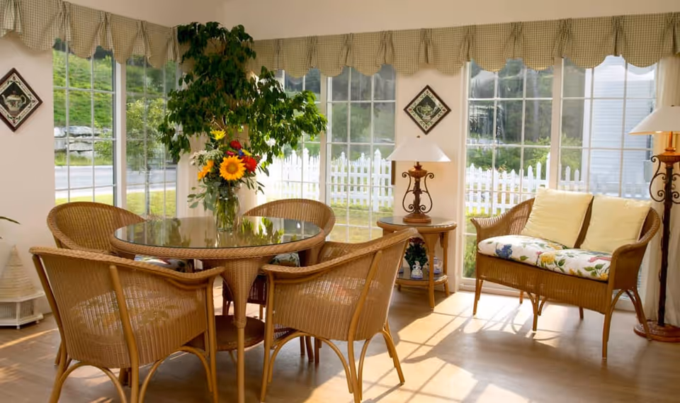 Sun-filled dining sunroom with a round glass-topped wicker table surrounded by wicker chairs, a cushioned wicker loveseat, lamps, and large windows showing a white picket fence.