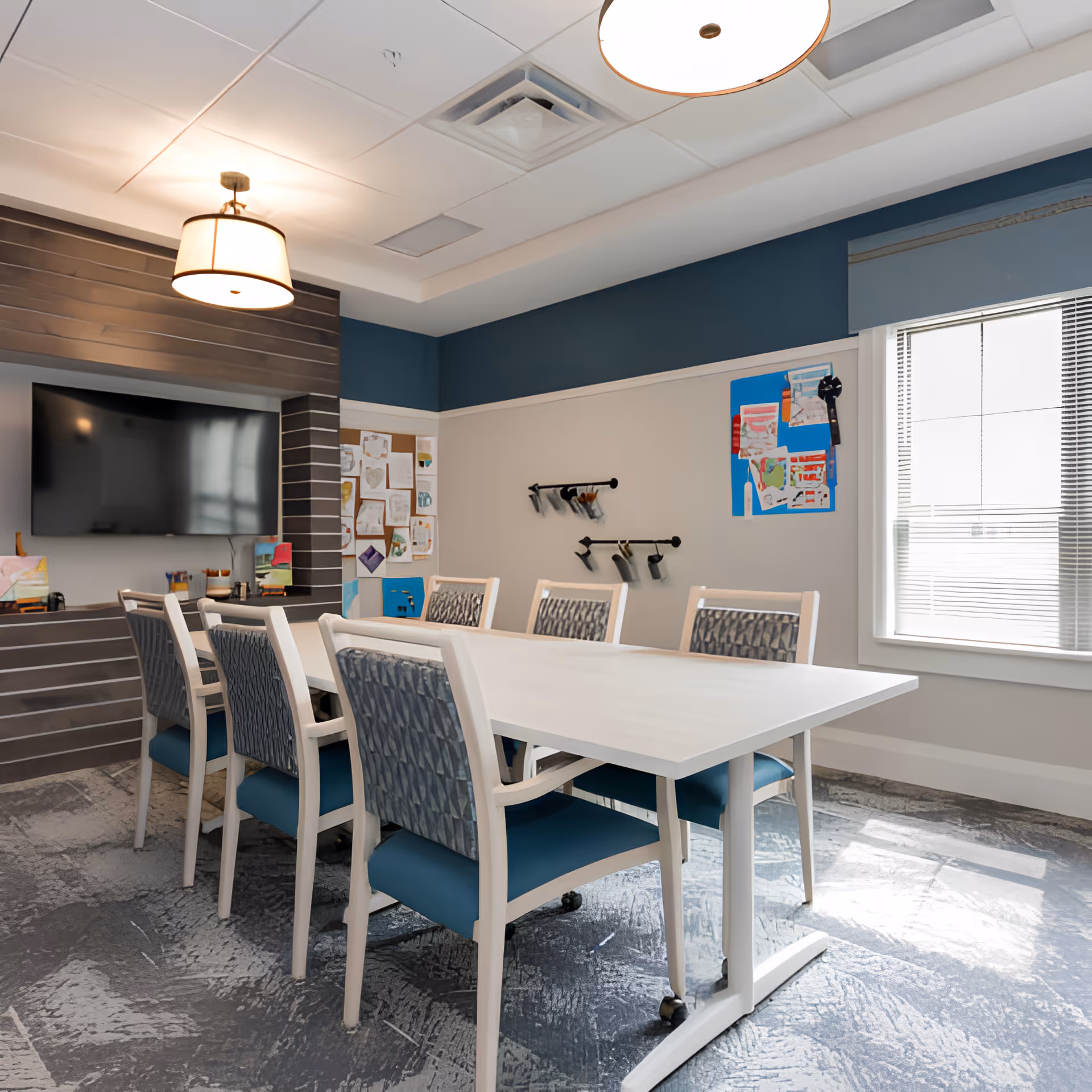 A well-lit meeting or activity room with a long white table surrounded by eight chairs with blue cushions and patterned backs. The room has a large flat-screen TV mounted on a wooden accent wall, bulletin boards with various papers and artwork, and a window with blinds letting in natural light. The ceiling has recessed lighting and a hanging light fixture.