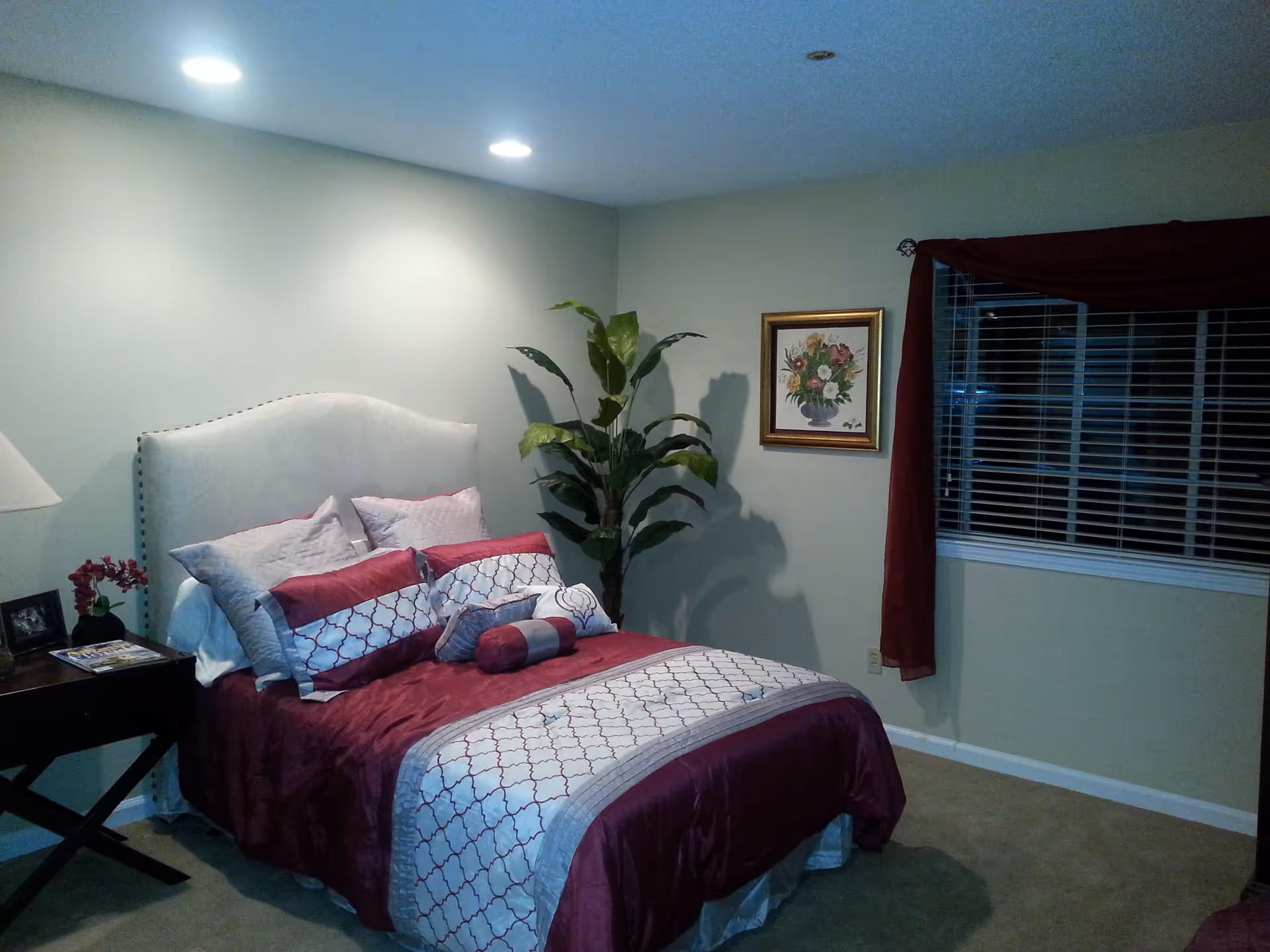 Bedroom with a double bed dressed in burgundy and white linens, a nightstand, potted plant, framed artwork, and a window with blinds.
