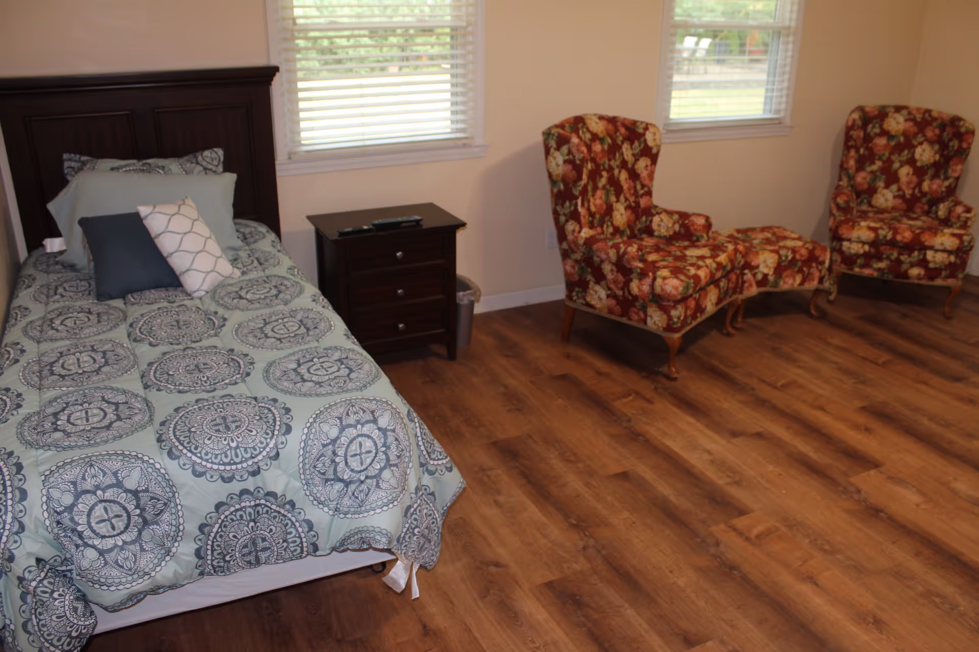 Bedroom with a single bed and nightstand, two floral armchairs and ottoman, and wood flooring under two windows.