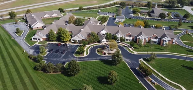 Aerial view of Heritage Pointe of Huntington, showing a large senior living facility with multiple connected buildings surrounded by well-maintained lawns, trees, and paved driveways with parking areas.
