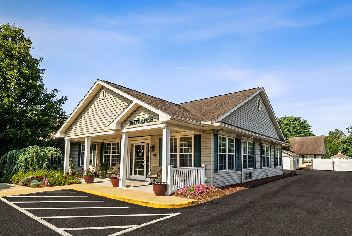 Exterior view of a single-story building with beige siding and a brown shingled roof, featuring a covered entrance with white columns and glass double doors. There are potted plants and chairs on the porch, a parking area with marked spaces, and trees and shrubs surrounding the building under a clear blue sky.
