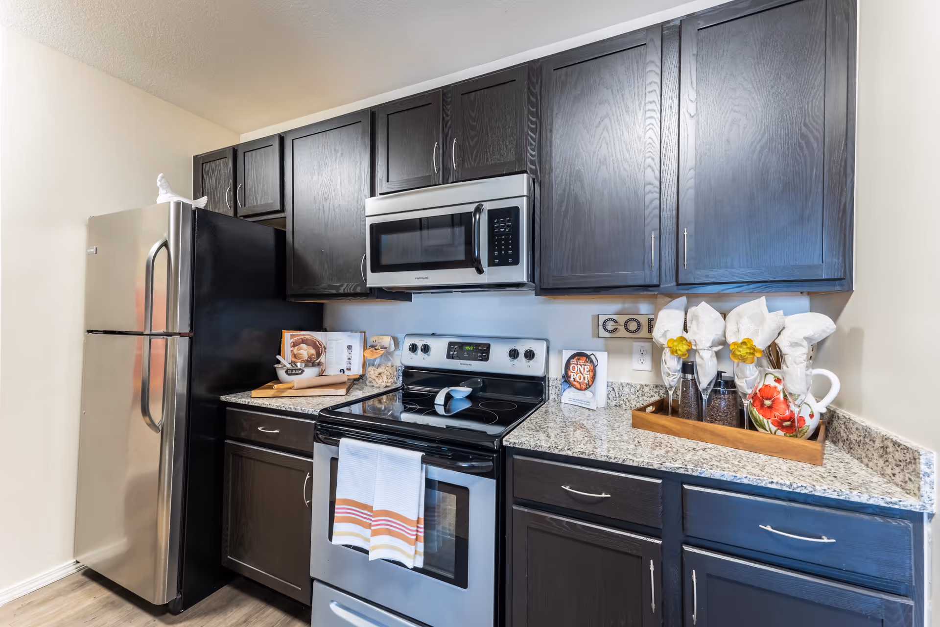 Modern kitchen with dark wood cabinets, stainless steel refrigerator, stove, and microwave. Granite countertops hold decorative items including a tray with glasses and napkins, a cookbook, and kitchen accessories.