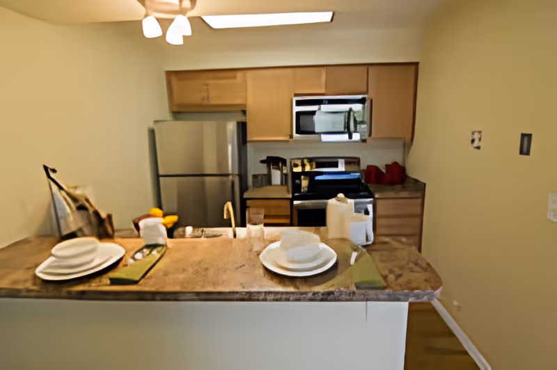 A small kitchen area with a countertop bar set for two with plates, bowls, and napkins. The kitchen features wooden cabinets, a stainless steel refrigerator, a microwave, and an oven. The walls are painted light yellow, and there is a ceiling fan with lights above.