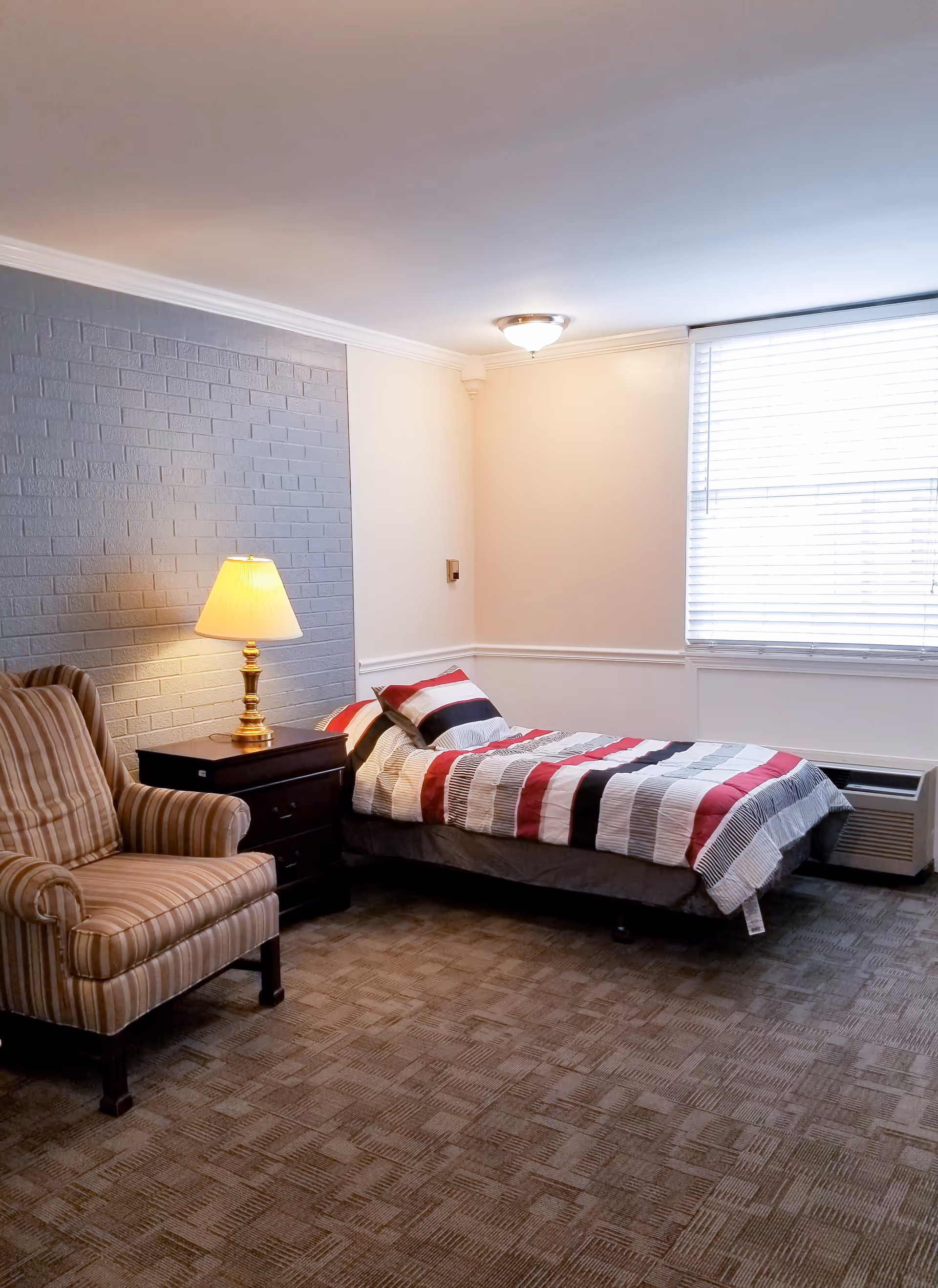 A senior living facility bedroom with a single bed covered in a striped red, black, white, and gray comforter. Next to the bed is a dark wooden nightstand with a brass lamp that is turned on. There is a striped armchair to the left of the nightstand. The walls are painted white with one accent wall in gray brick. A window with white blinds is on the right side of the room, and the floor is carpeted in a patterned brown design.