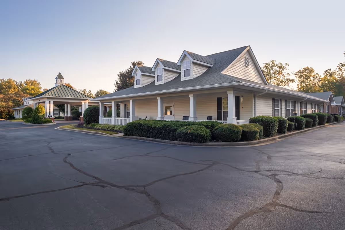 Exterior view of a single-story assisted living building with a covered porch, trimmed bushes, and a paved driveway.