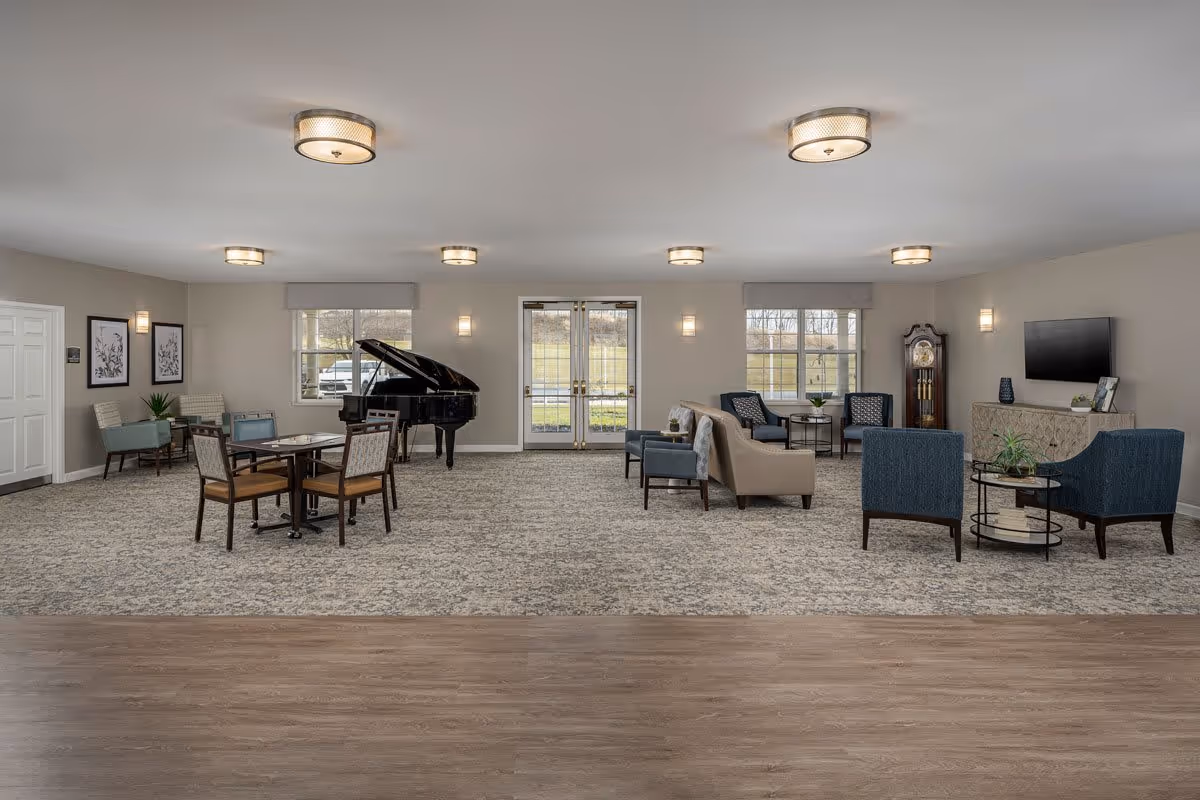 A spacious senior living facility common area with a mix of seating options including armchairs, a sofa, and a table with chairs. There is a grand piano near the windows and a grandfather clock against the wall. The room has large windows and double glass doors letting in natural light, neutral-colored walls, and a combination of carpet and wood flooring.