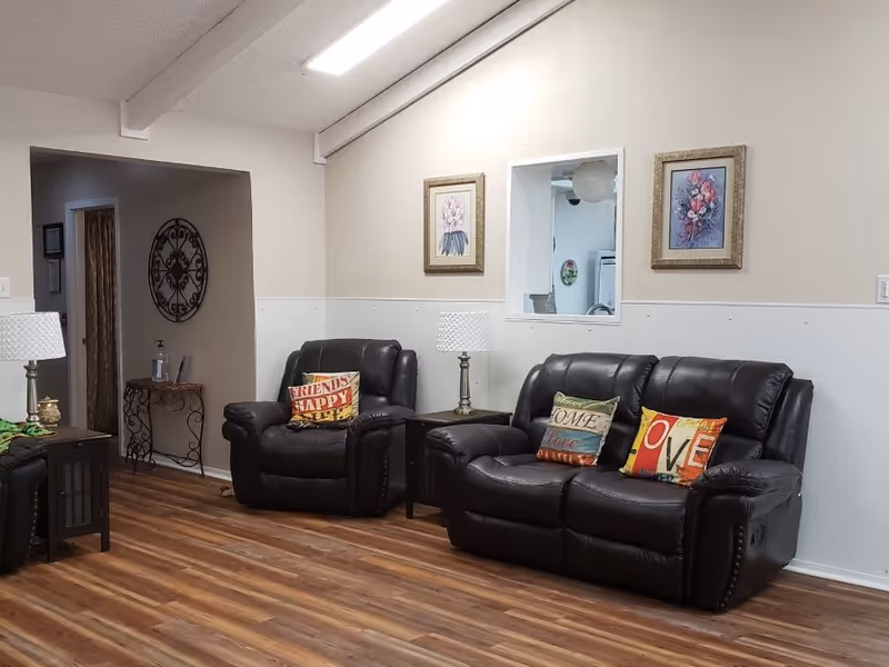 Cozy living room with black leather recliners and a loveseat, colorful throw pillows, side tables with lamps, framed artwork, and wood-look flooring.