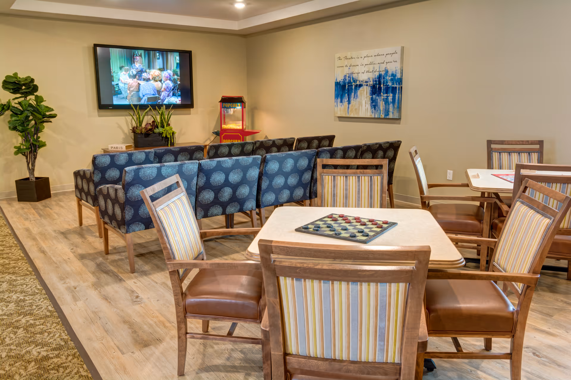 A cozy common area with several wooden tables and chairs featuring striped upholstery. A checkers game board is placed on one of the tables. In the background, there are blue patterned armchairs arranged facing a wall-mounted TV showing a group of people. A popcorn machine and a potted plant are also visible, along with a blue and white abstract painting on the wall.