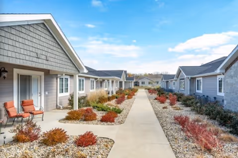 Central walkway flanked by single-story assisted-living units with rock landscaping and patio chairs under a blue sky.