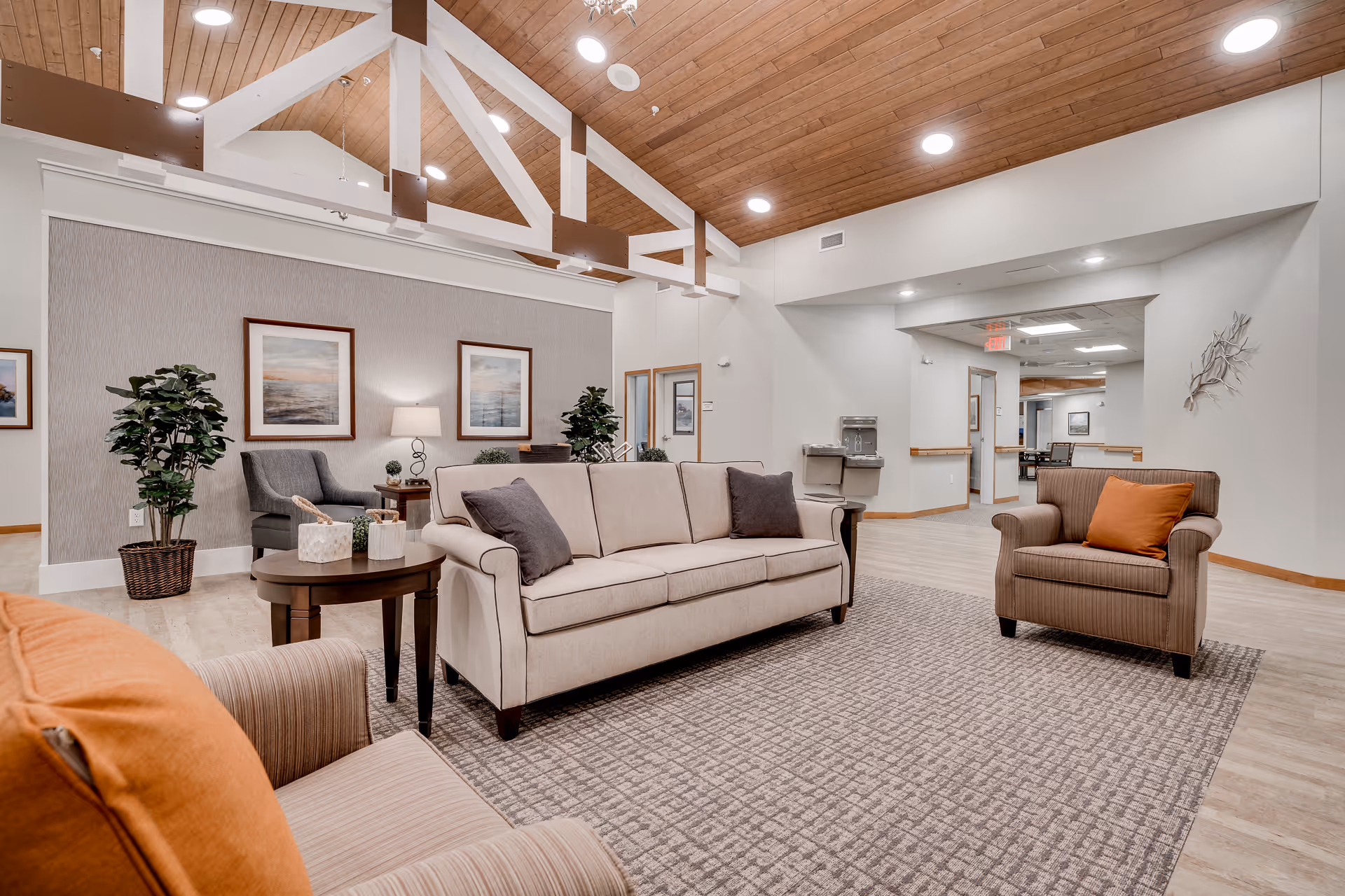 A spacious and well-lit senior living facility common area with a high wooden ceiling and exposed beams. The room features a beige sofa with two dark gray pillows, a striped armchair with an orange pillow, a round wooden side table with decorative items, and a gray armchair near the back wall. The walls are decorated with framed artwork, and there are potted plants adding greenery. The floor is covered with a patterned area rug and light wood flooring. In the background, there is a water fountain and a hallway leading to other rooms.