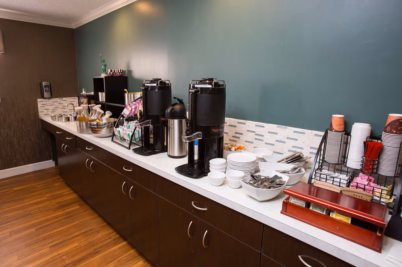 A coffee and tea station with two large black coffee dispensers, a stainless steel airpot, stacks of white bowls and plates, utensils in white bowls, a rack with coffee cups and stirrers, and various tea packets on a long countertop with dark brown cabinets below and a teal wall with a tiled backsplash behind.