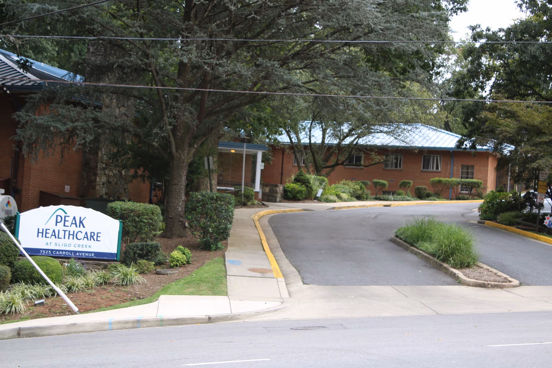 Exterior view of a healthcare facility named Peak Healthcare at Sligo Creek, showing a driveway, sidewalk, trees, shrubs, and a brick building with a blue roof in the background.
