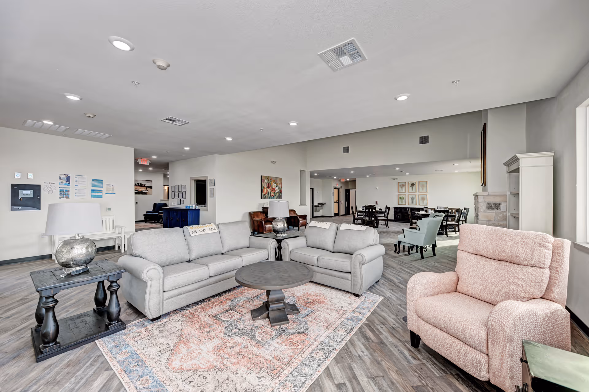 Spacious assisted-living common lounge with sofas, armchairs, a coffee table, and a dining area visible in the background.