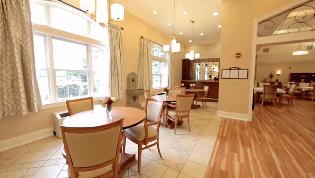 A bright dining room area with round wooden tables and cushioned chairs. Large windows with patterned curtains allow natural light to fill the space. There are pendant lights hanging from the ceiling and a small decorative fountain near the windows. The room has a combination of tiled and wooden flooring, and an adjoining room with more tables and chairs is visible.