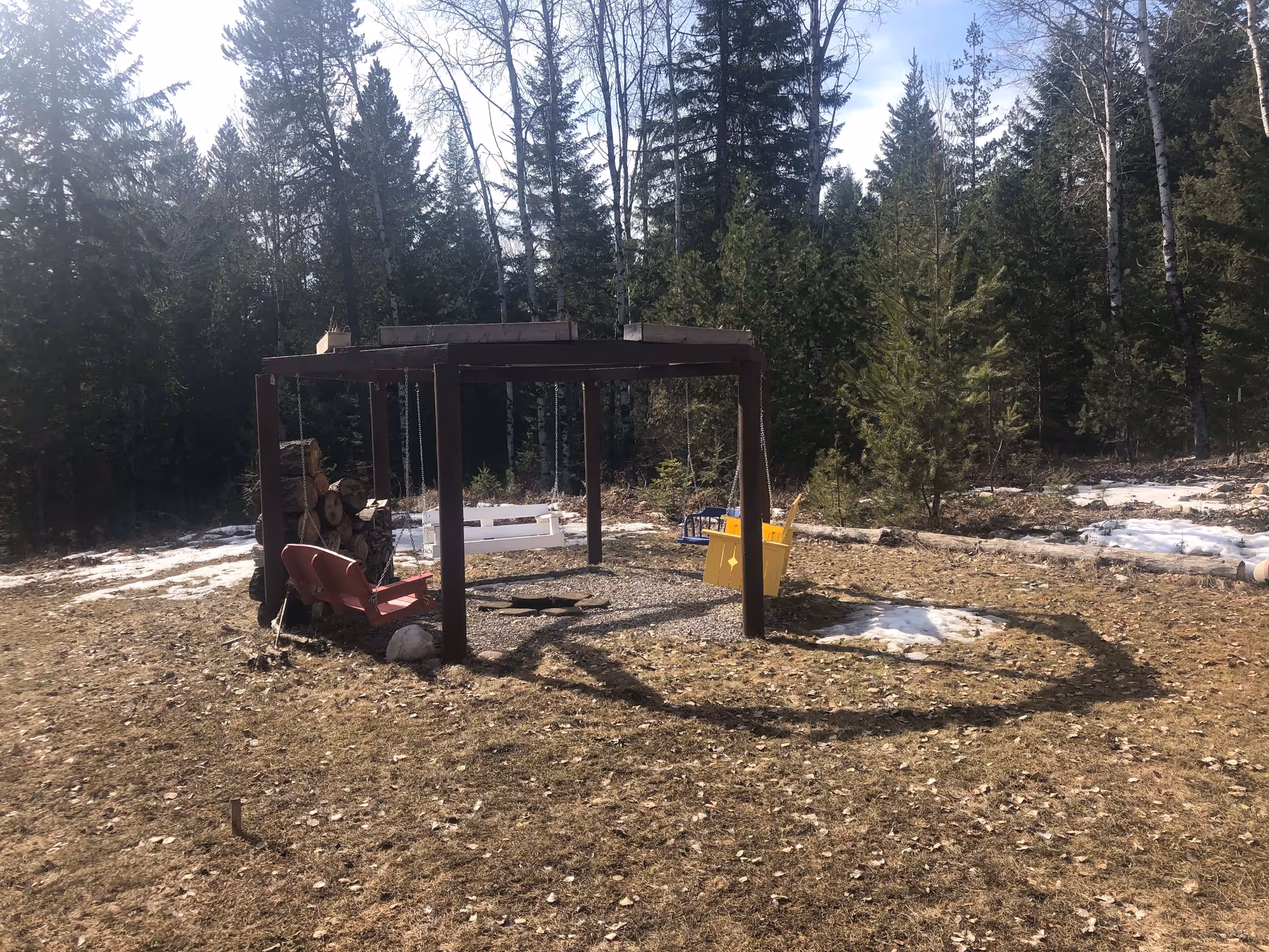 Outdoor seating area with a wooden pergola structure supporting four colorful swings in red, white, blue, and yellow. The area is surrounded by a forest with tall evergreen trees and patches of snow on the ground.