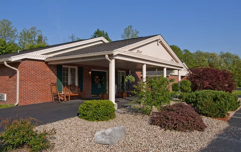 Exterior view of a single-story brick building with a covered porch supported by white columns. The porch has wooden rocking chairs and potted plants. The surrounding landscape includes various bushes, shrubs, and a rock garden under a clear blue sky.