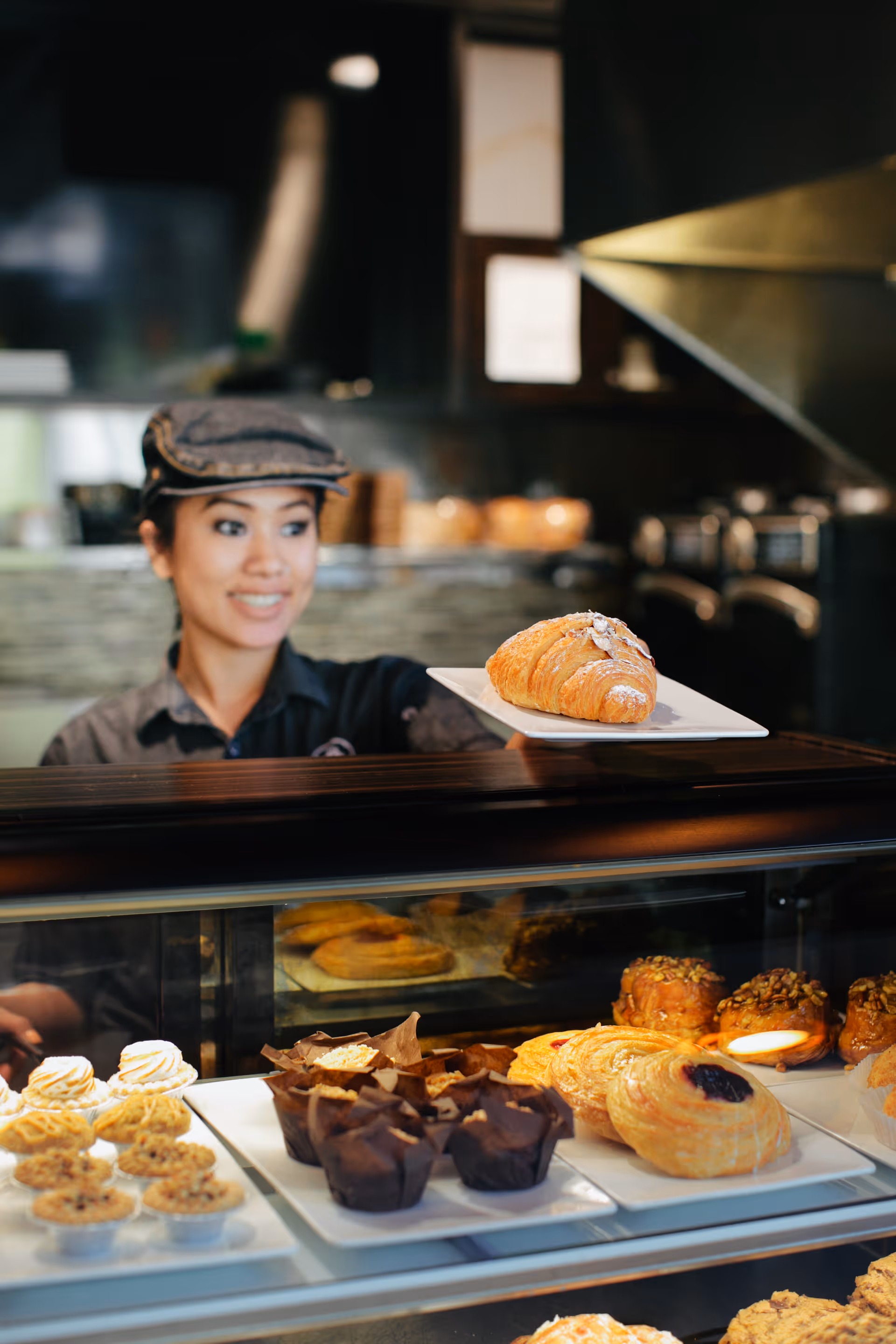 A smiling woman wearing a cap and uniform behind a bakery display case, holding a plate with a croissant. Various pastries and baked goods are visible inside the glass display case.