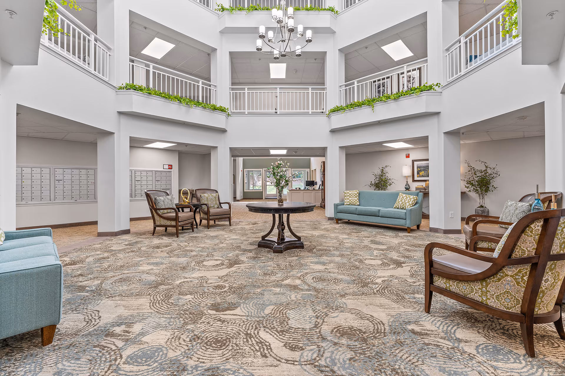 Spacious and well-lit lobby area of Jordan Oaks Gracious Retirement Living featuring a patterned carpet, multiple seating arrangements with sofas and armchairs, a central round table with a floral arrangement, white railings on the upper level with green plants, and mailboxes along the walls.