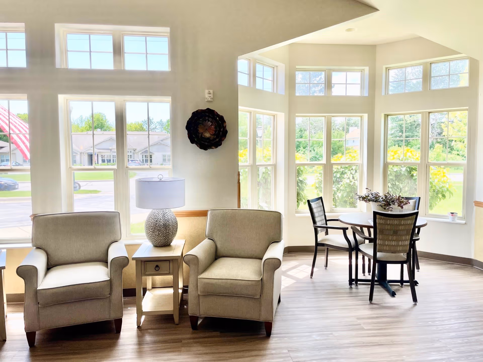 A bright and airy sitting area with two beige armchairs separated by a small wooden side table with a decorative lamp. In the background, there is a round table with four chairs near large windows that let in natural light and offer a view of greenery outside.