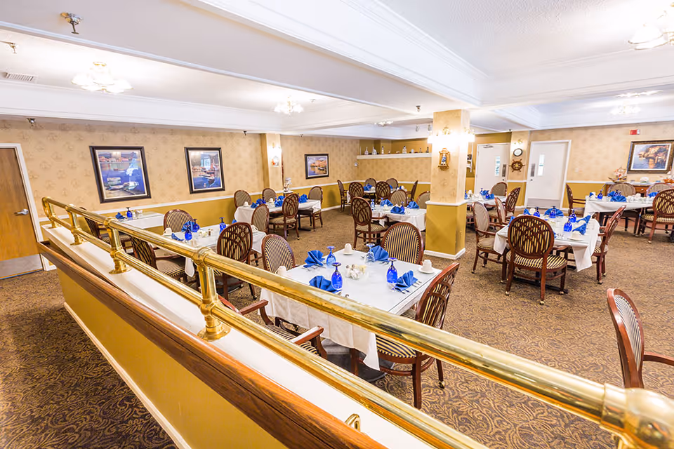 A spacious dining room with multiple tables covered in white tablecloths, each set with blue napkins, blue glasses, and white cups. The room has patterned carpet, wallpapered walls with framed artwork, and brass railings along a raised platform area. Wooden chairs with striped upholstery surround the tables, and ceiling lights illuminate the space.