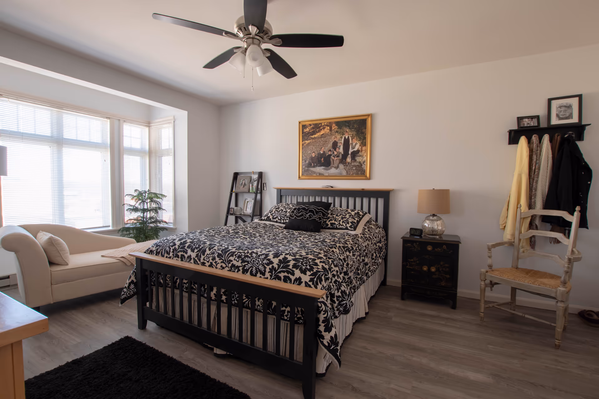 A well-lit bedroom with a large bed featuring a black and white floral patterned comforter. There is a beige chaise lounge near a large window with blinds, a black nightstand with a lamp, a wooden chair, and a wall-mounted coat rack with clothes hanging. A ceiling fan with lights is mounted on the white ceiling, and a framed family photo hangs above the bed.