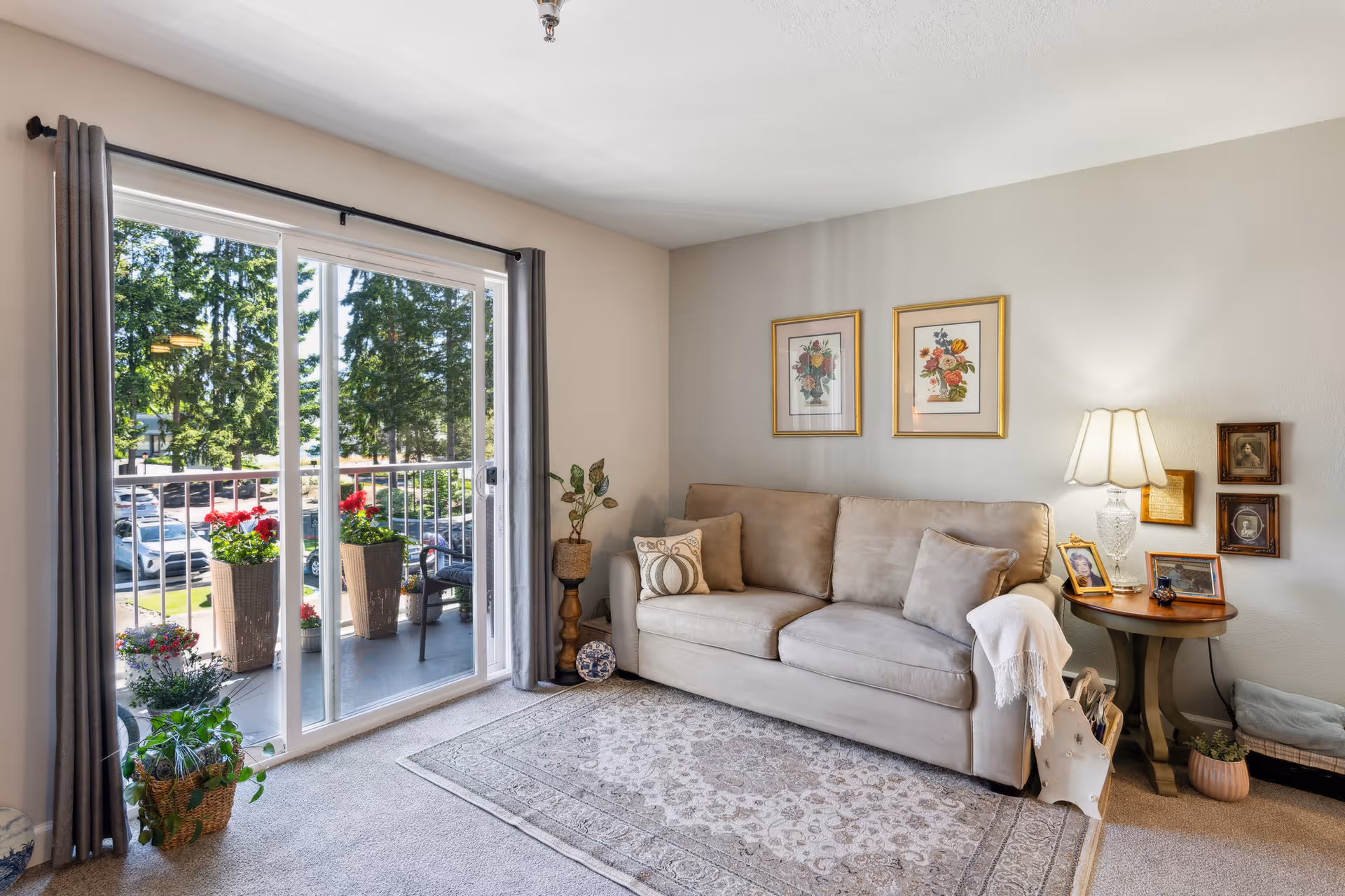 Bright living room with a beige sofa, side table and lamp, area rug, and sliding glass doors opening to a balcony with potted plants.