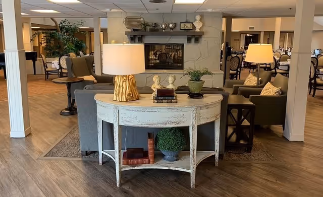 A cozy senior living common area with a white distressed console table in the foreground holding a lamp, two bust sculptures, books, and a potted plant. Behind the table are comfortable armchairs and sofas arranged around a fireplace with decorative items on the mantel. In the background, there are dining tables and chairs, and the space has wooden flooring and warm lighting.